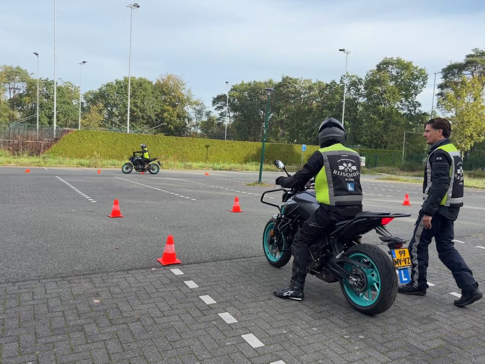 Leerling op de lesmotor tussen de pionnen op het oefenterrein, de instructeur loopt mee