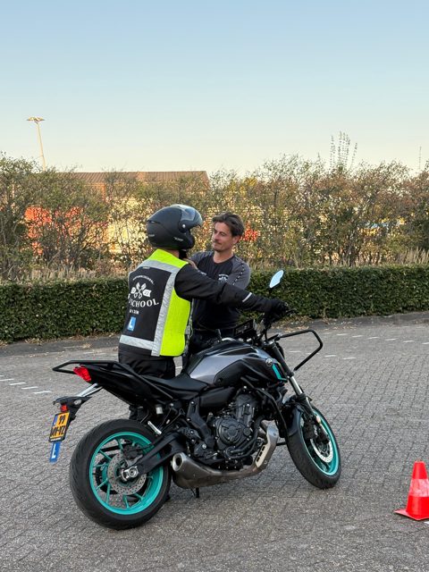 Instructeur helpt een leerling met een geel hesje op de lesmotor, op het oefenterrein
