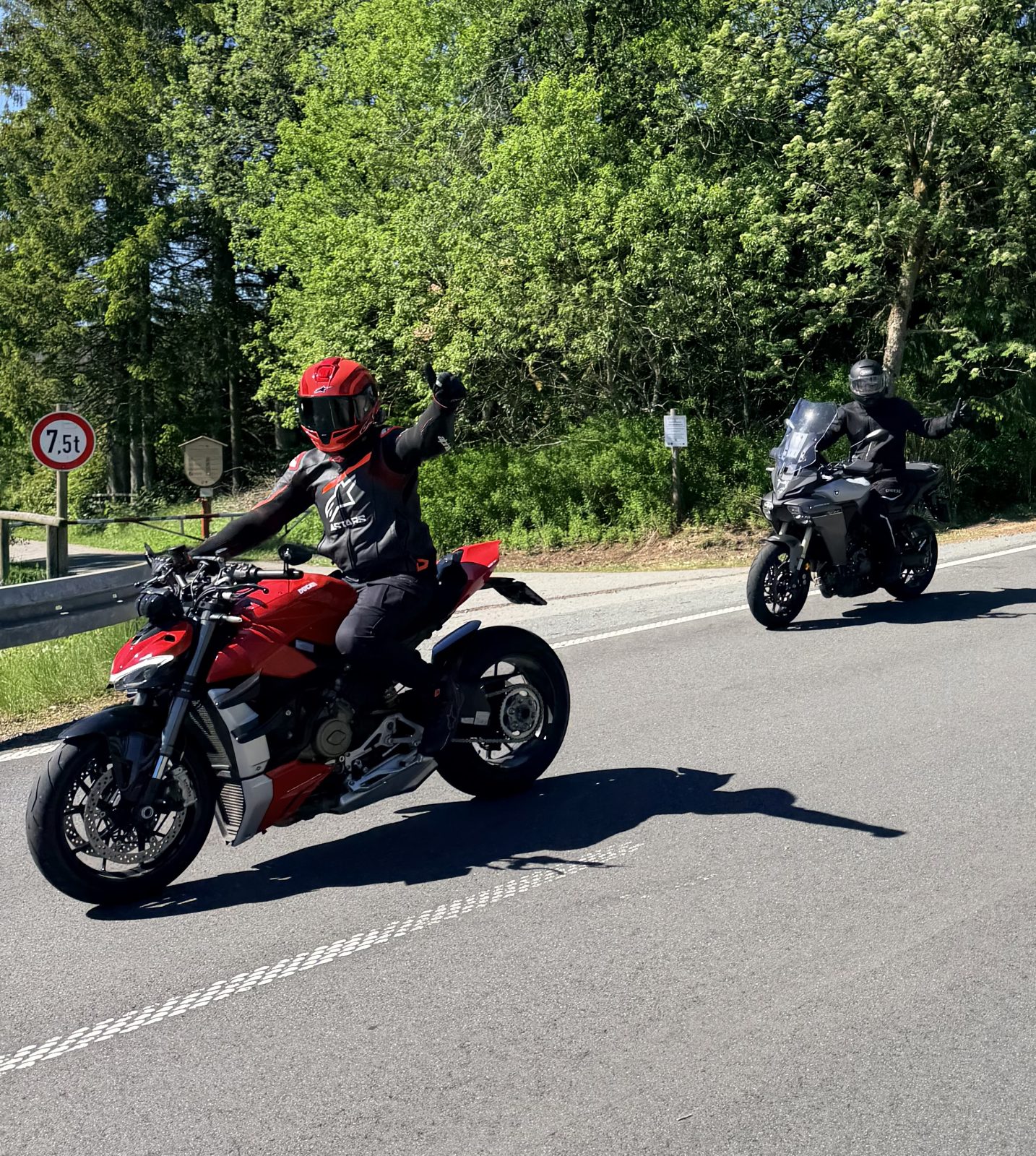 Twee motorrijders zwaaien vanaf hun motor op een weg langs het bos, voorop een rode motor