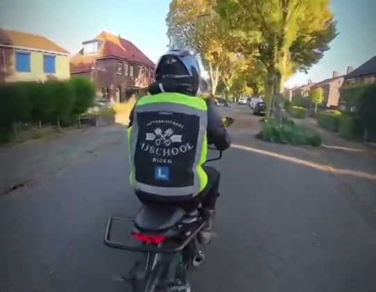 Leerling in het hesje van Motorrijschool Rijen rijdt door een woonwijk, gefilmd vanaf de motor van de instructeur
